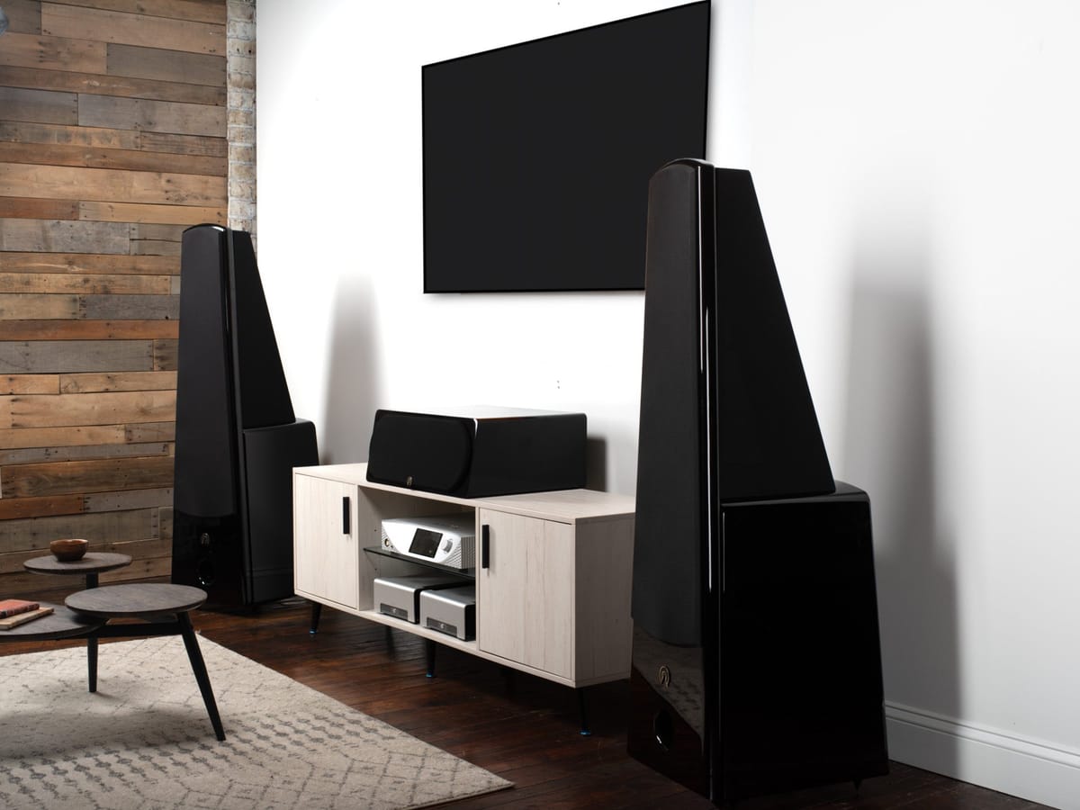 A pair of Alta Audio Aphrodite loudspeakers either side of a wall mounted screen in a loft living room with a reclaimed timber wall and dark hardwood floor